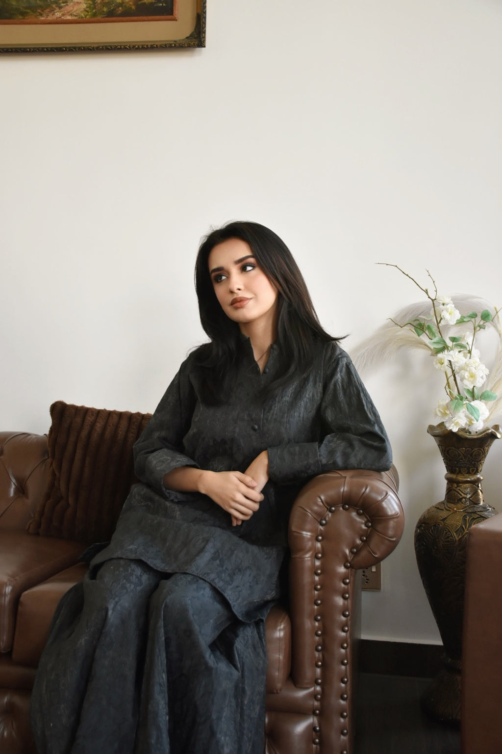 Woman sitting on a brown leather couch in a room with a white wall and decorative elements.